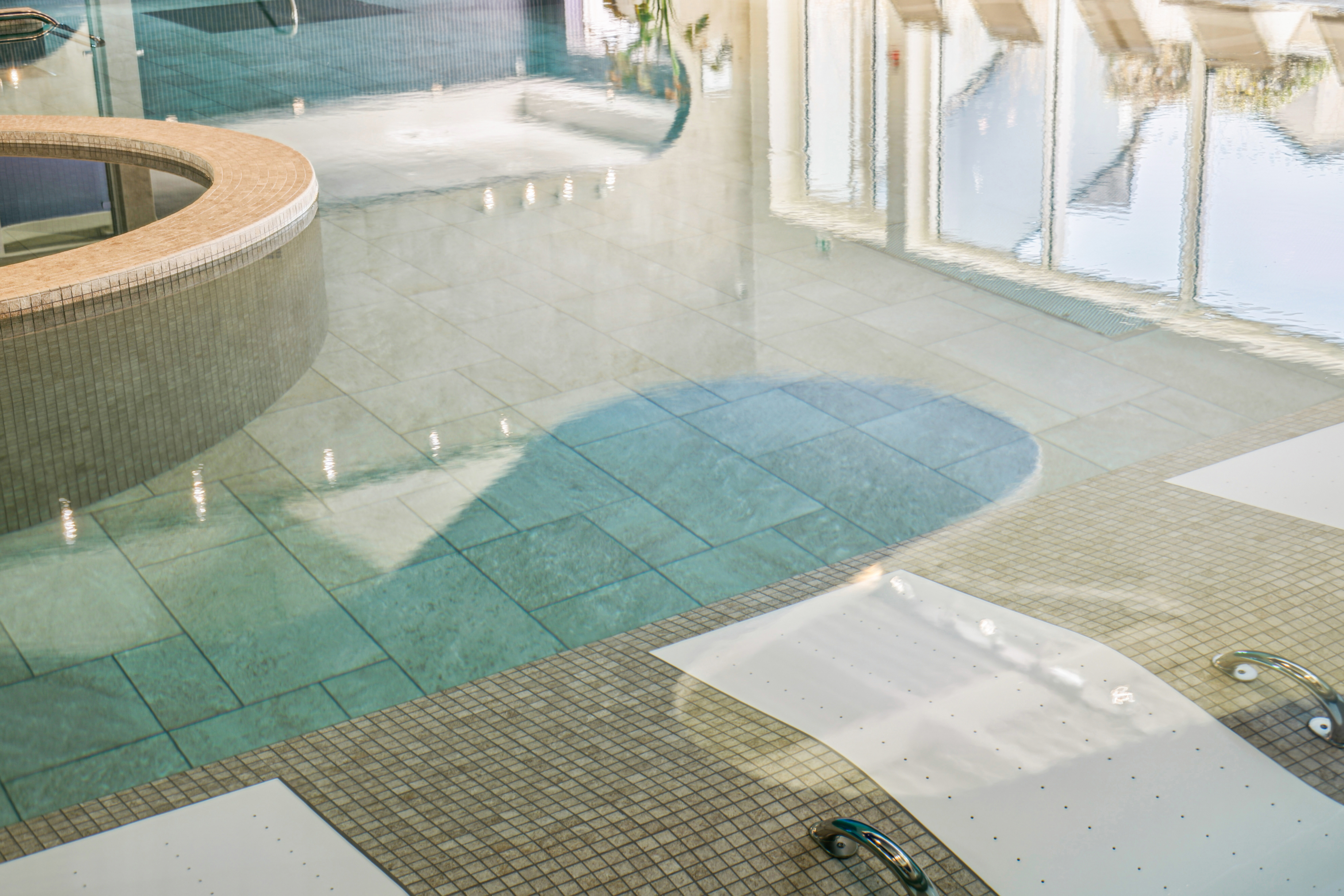Vue rapprochée de la piscine intérieure montrant des lits de relaxation immergés, dallage clair et reflets des baies vitrées sur l’eau.