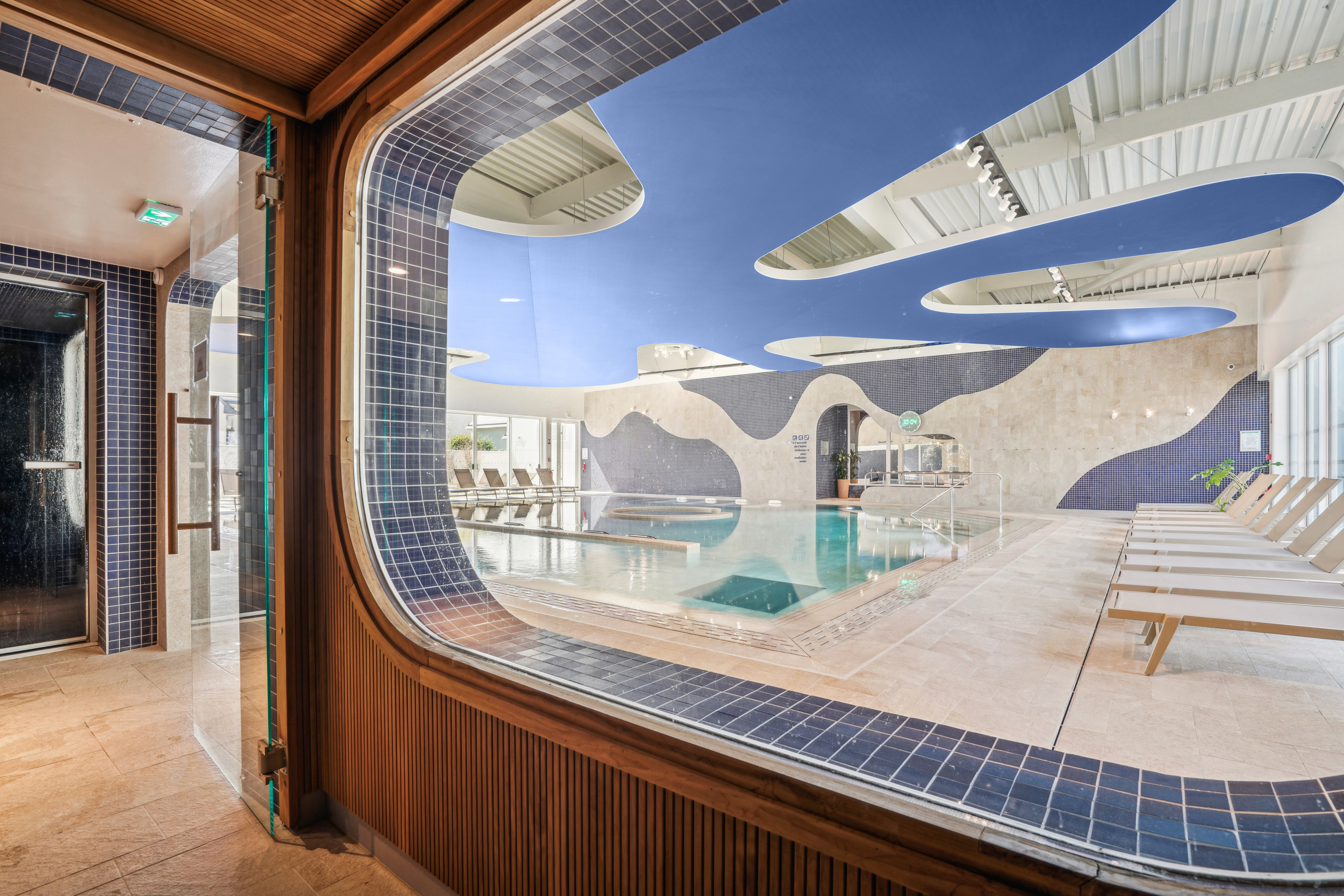 Espace bien‑être intérieur avec grande piscine, transats alignés et plafond acoustique bleu aux formes organiques, vu à travers une large vitre encadrée de mosaïques bleues.