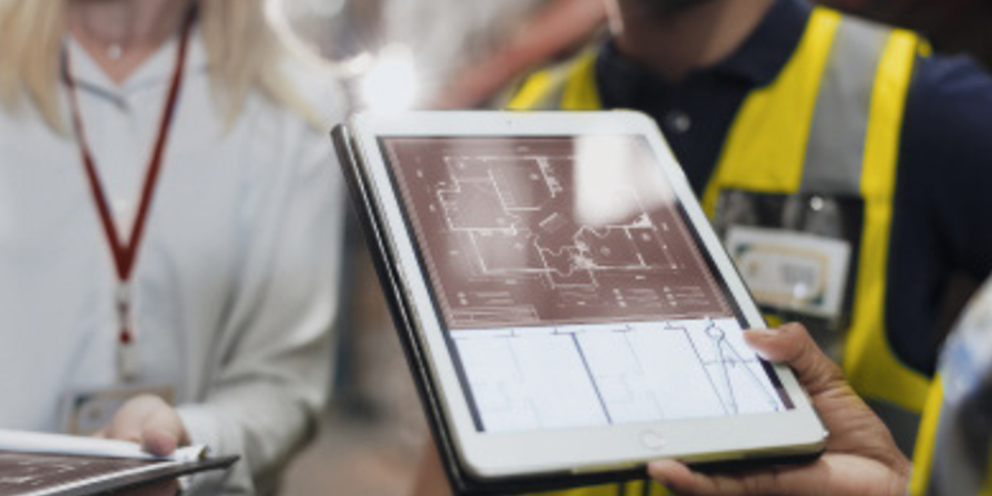 Plusieurs personnes, dont des professionnels en gilets haute visibilité, consultent un plan architectural affiché sur une tablette numérique. La scène se déroule dans un environnement industriel ou logistique, et les participants semblent échanger autour de la configuration ou de la conception d’un espace.