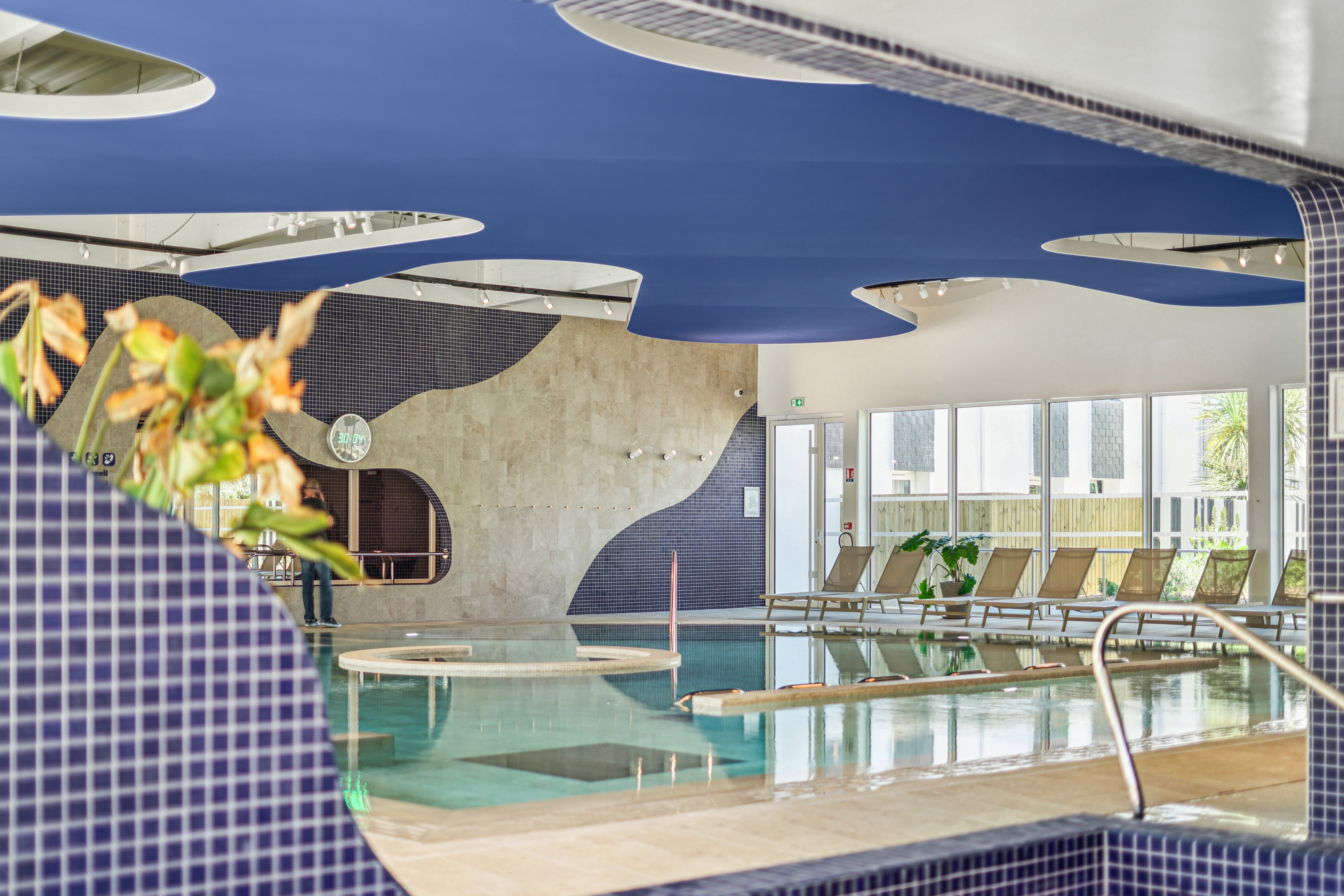 Plafond bleu aux courbes fluides, inspiré d’un splash d’eau, surplombant la piscine et dialoguant avec les murs carrelés bleu marine du spa.