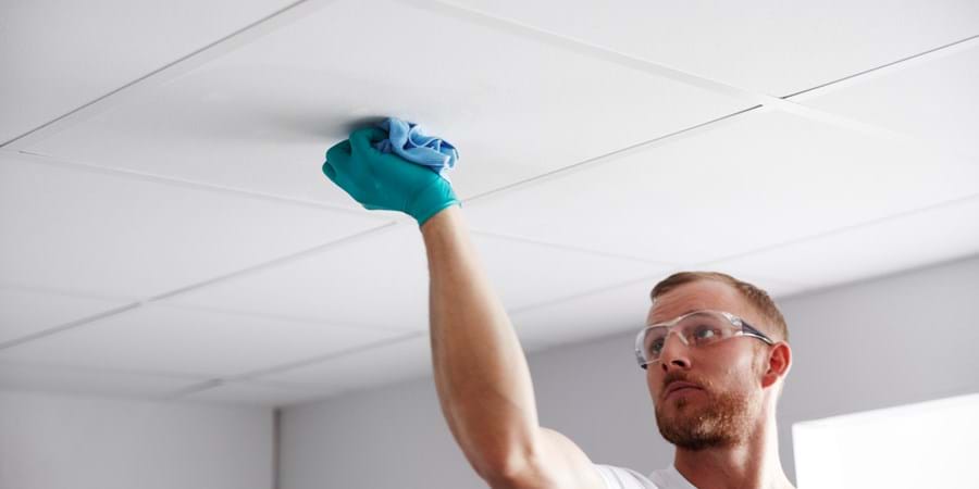 A man wearing safety glasses and a blue glove cleans a white ceiling tile with a cloth in a brightly lit room.