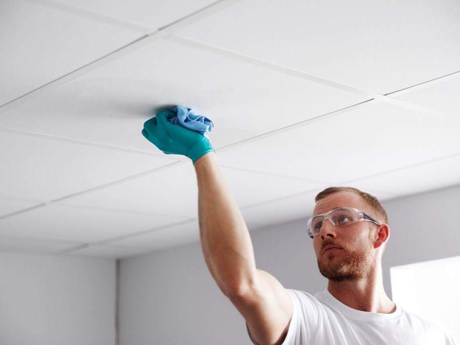 A man wearing safety glasses and a blue glove cleans a white ceiling tile with a cloth in a brightly lit room.