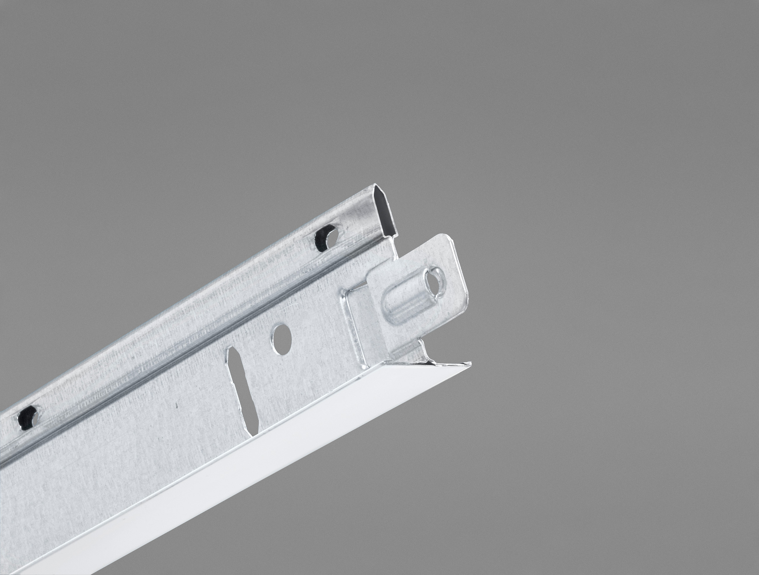 Primo piano di una griglia metallica per controsoffitto sospeso (T-bar) su uno sfondo grigio uniforme, che mostra fori e scanalature per l’installazione. La T-bar presenta una finitura metallica e bordi puliti e lineari.