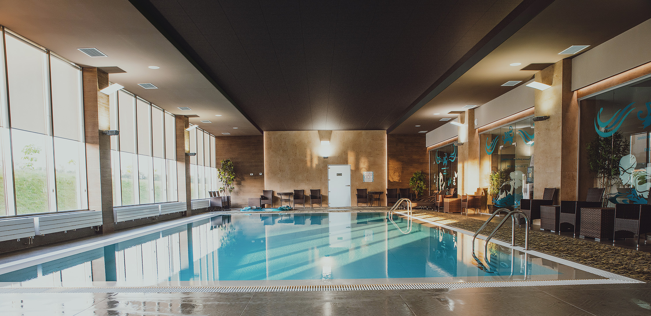 A swimming pool area with soft, natural colours on the floor and walls. Black acoustic panels are installed in the ceiling. To the left there are big windows bringing natural light into the room. Dark brown rattan chairs are placed along the walls and green plants are seen in the corners of the room.