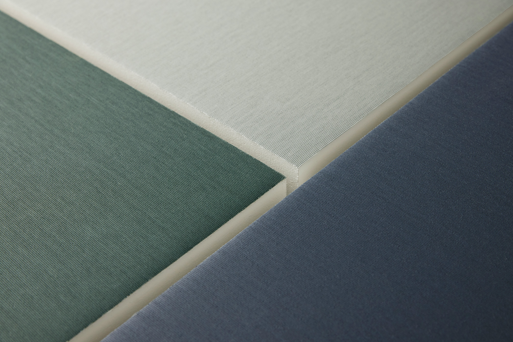 Close-up of three fabric-covered acoustic panels in soft green, light grey, and deep blue tones, showing their textured surfaces and the sharp, angled edges where the panels meet.