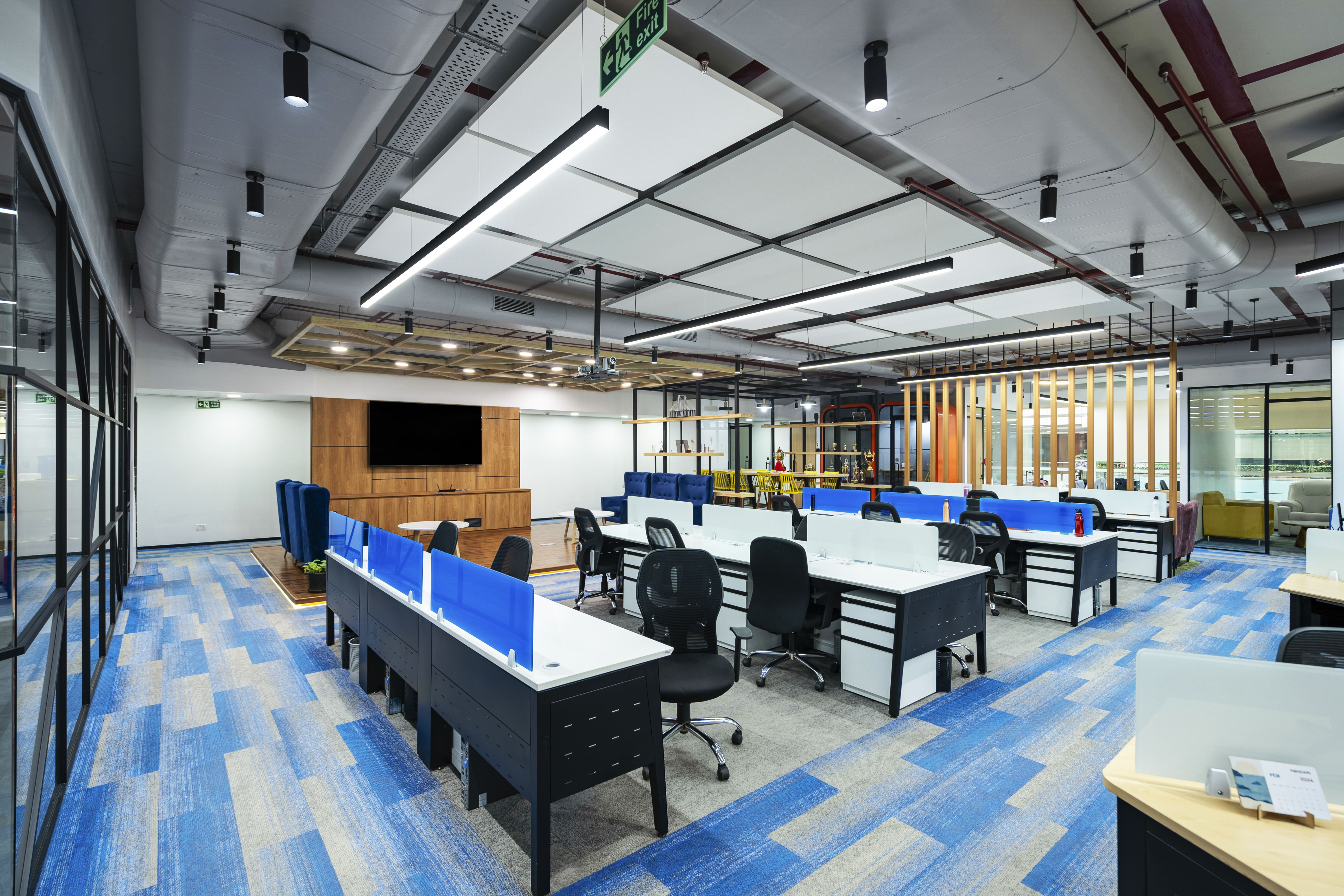 An empty modern office space with blue carpeting. The area is well-lit with overhead lights and features a combination of cubicles with blue partitions and black office chairs. To the left, there is a wooden panel wall with a large screen, and in the background, there are wooden pillars. Visible are also air ducts and free-hanging square acoustic panels in white and grey forming a pattern in the ceiling.