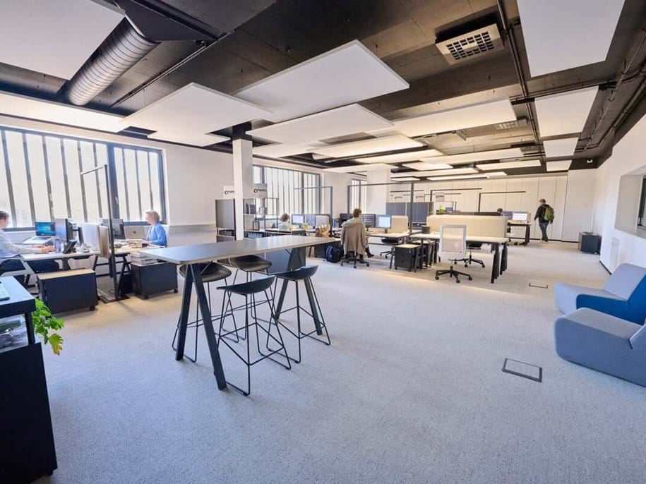A modern open-plan office with several people working at desks, computers, and monitors. There are tall tables with bar stools, large windows letting in natural light, and contemporary office furniture.