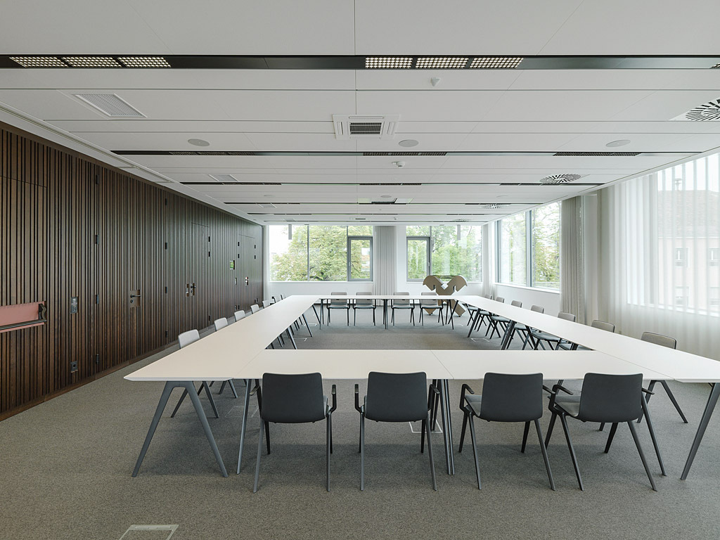 Suspended, acoustic ceiling in large meeting room with a rectangular seating arrangement, a dark wooden wall and white blinds covering part of the big windows.