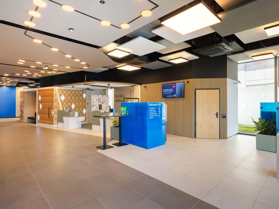 Modern bank interior with bright lighting, tiled flooring, blue service counters, digital screens, and wooden accents. Glass-walled offices and waiting areas are visible, creating an open, welcoming space.