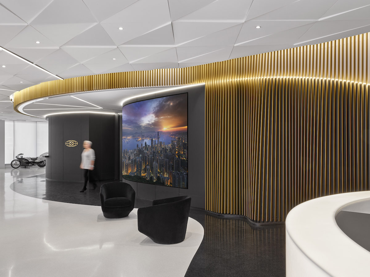 Modern office lobby with sleek design features a large curved wall with vertical gold paneling and an embedded screen showing a cityscape. Two black chairs are in front of the screen, and a person is blurred walking by. A motorcycle is in the background.