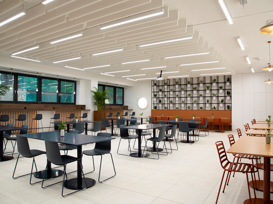 A modern, spacious canteen with black and red chairs, black tables, large windows, ceiling lights, and a decorative wall with plants. The space is bright, tidy, and features contemporary design elements.