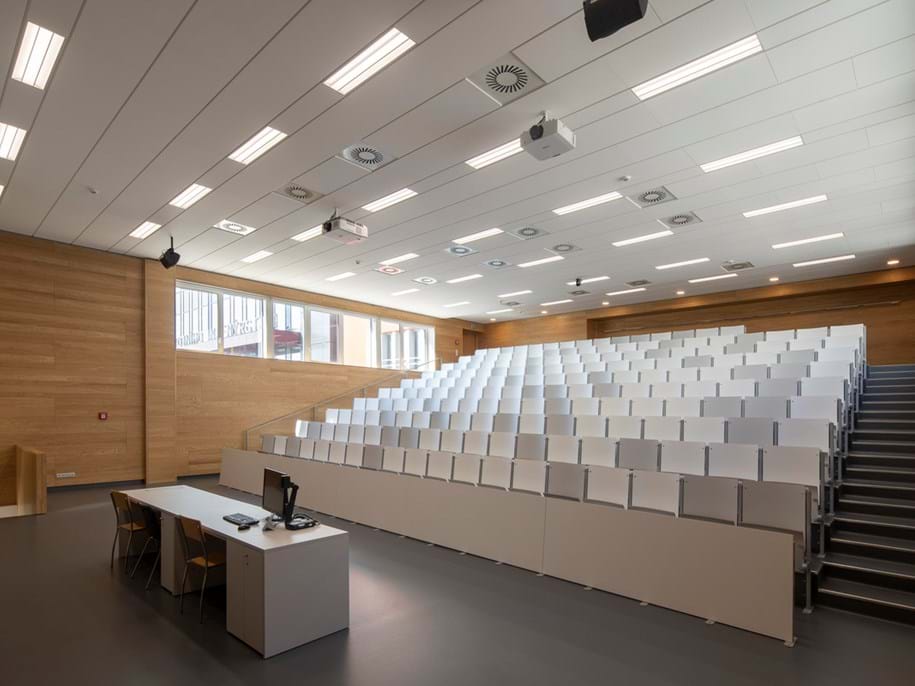 White, suspended acoustic ceiling in lecture hall with wooden walls and a slanting floor with rows of chairs