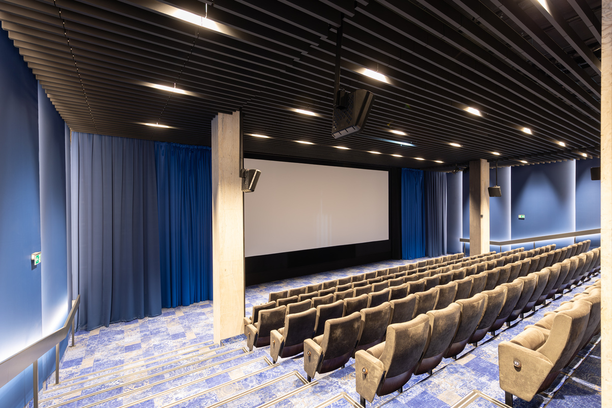 A large cinema with rows of upholstered chairs and a big screen with blue curtains on the sides. Acoustic baffles are installed in the ceiling. The walls are blue and the carpet has a matching blue and beige pattern.