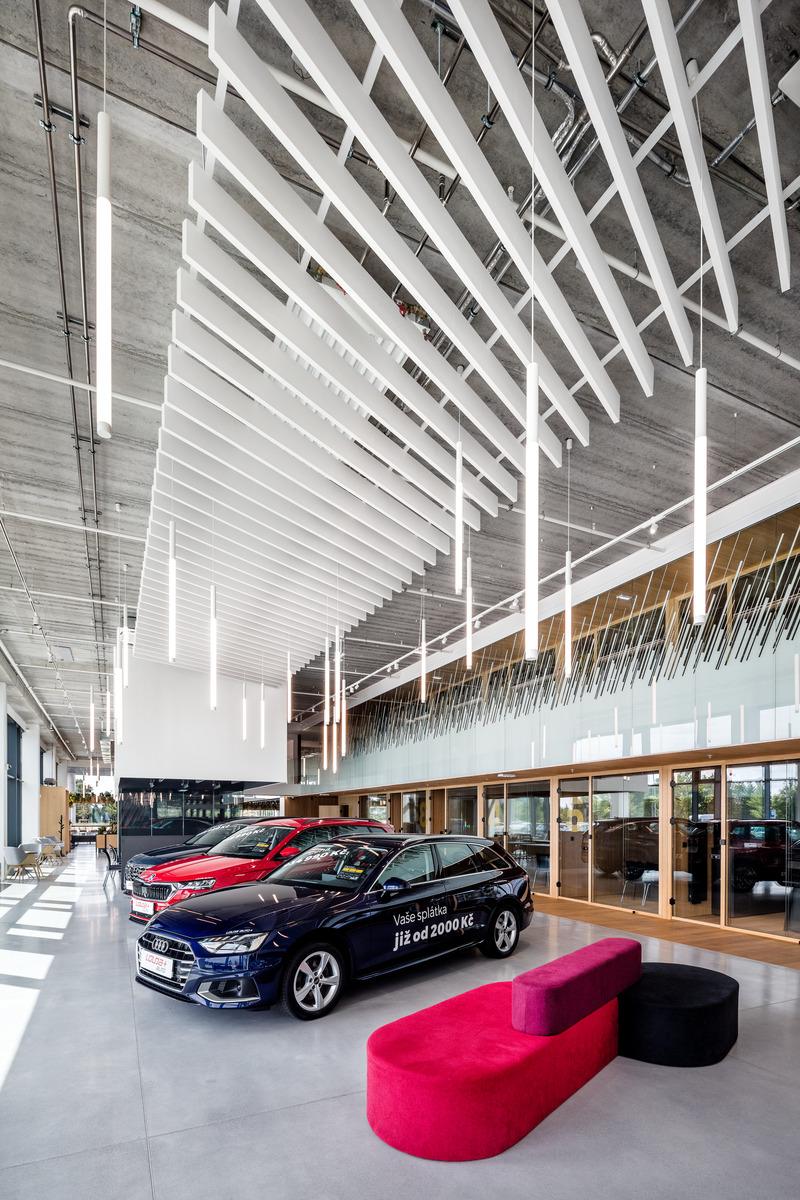 Interior of a modern car showroom with high ceilings featuring an array of white accoustic baffles. Three cars are on display, one in the foreground and two slightly behind it to the left. The floor is polished concrete, and large windows allow natural light to fill the space. In the centre, there is a red rectangular seating area with two round black stools. The showroom appears spacious and designed to highlight the vehicles for sale.