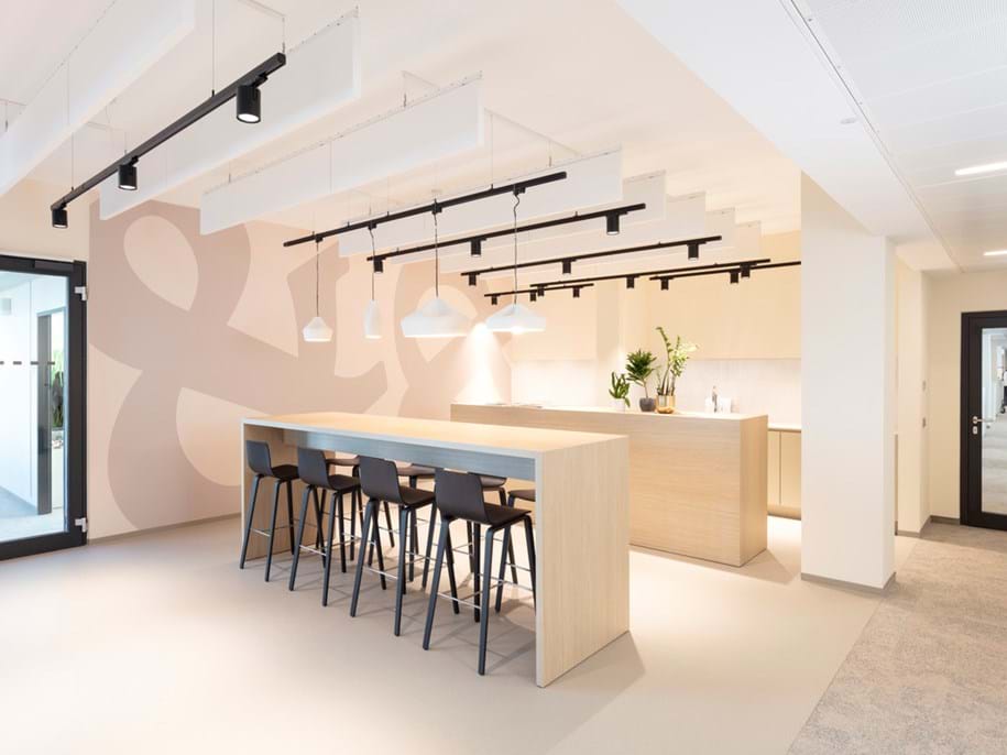 A modern office kitchen with a long wooden island, black bar stools, pendant lighting, and minimalistic decor. A large ampersand design is on the wall, and there are plants on the counter. Glass doors lead to adjoining rooms.