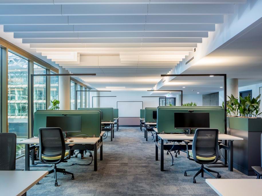 Modern open-plan office with rows of green desk partitions, ergonomic chairs, computer monitors, large windows on the left, and ceiling panels overhead. Potted plants add greenery to the bright, spacious workspace.