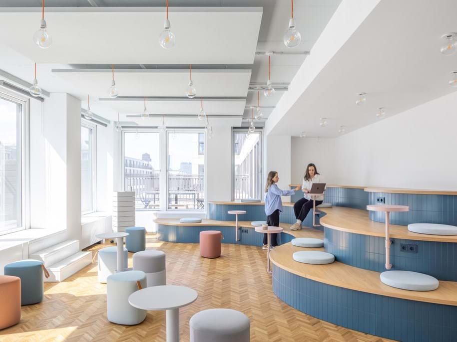 A modern, bright office lounge with tiered seating, round pouffes, large windows, and two women talking whilst one uses a laptop. Light wood floors and white walls create an open, airy atmosphere.