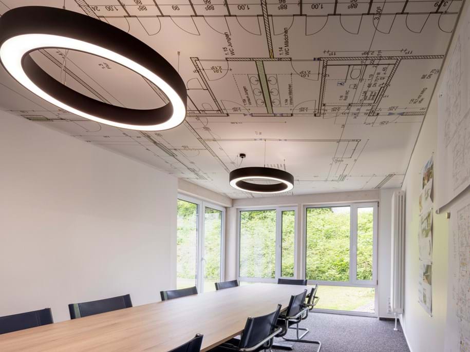 A modern conference room with a long wooden table, black chairs, large windows showing trees outside, and two circular ceiling lights; architectural floor plans are printed on the ceiling.