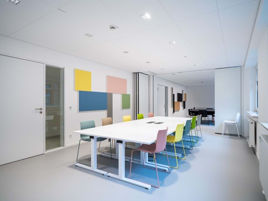 A modern meeting room with white walls, colourful square wall panels, and a long white table surrounded by pastel-coloured chairs. Large windows on the right let in natural light.