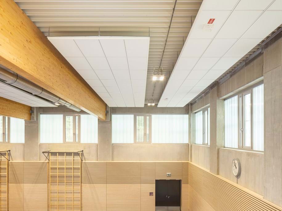 A modern gymnasium interior with light wood and concrete walls, large windows, wall bars, exposed beams, a clock, and a high ceiling with white acoustic panels.