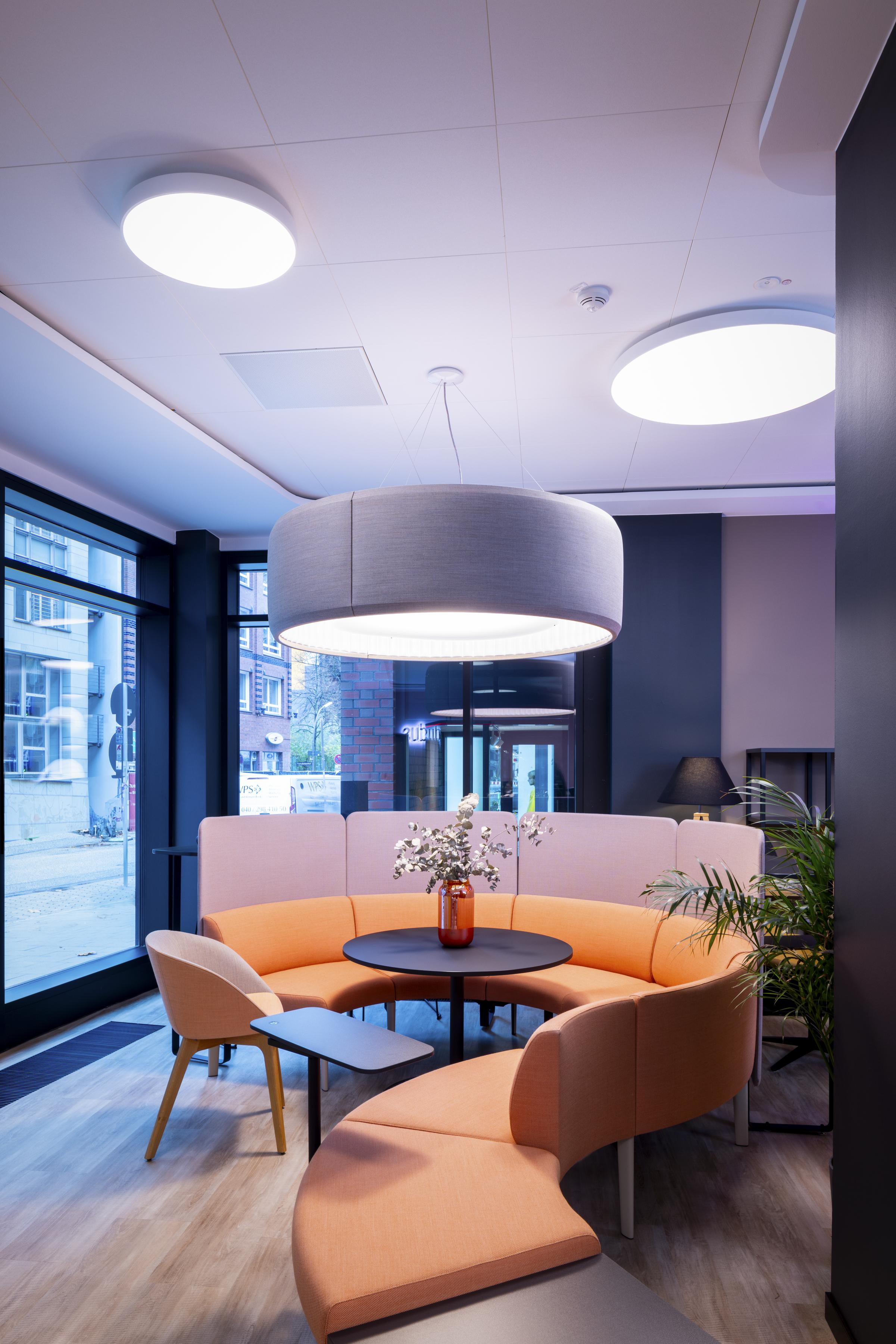 White, suspended acoustic ceiling, a swirling orange sofa and big, grey ceiling lamp in office furnture showroom 