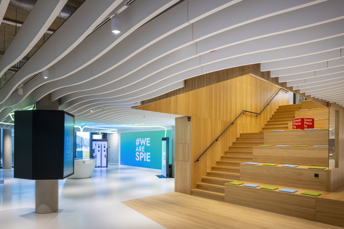 A modern office interior with wavy ceiling panels, wooden tiered seating with colourful cushions, and a large turquoise wall displaying the text #WE ARE SPIE. The space is brightly lit and open.