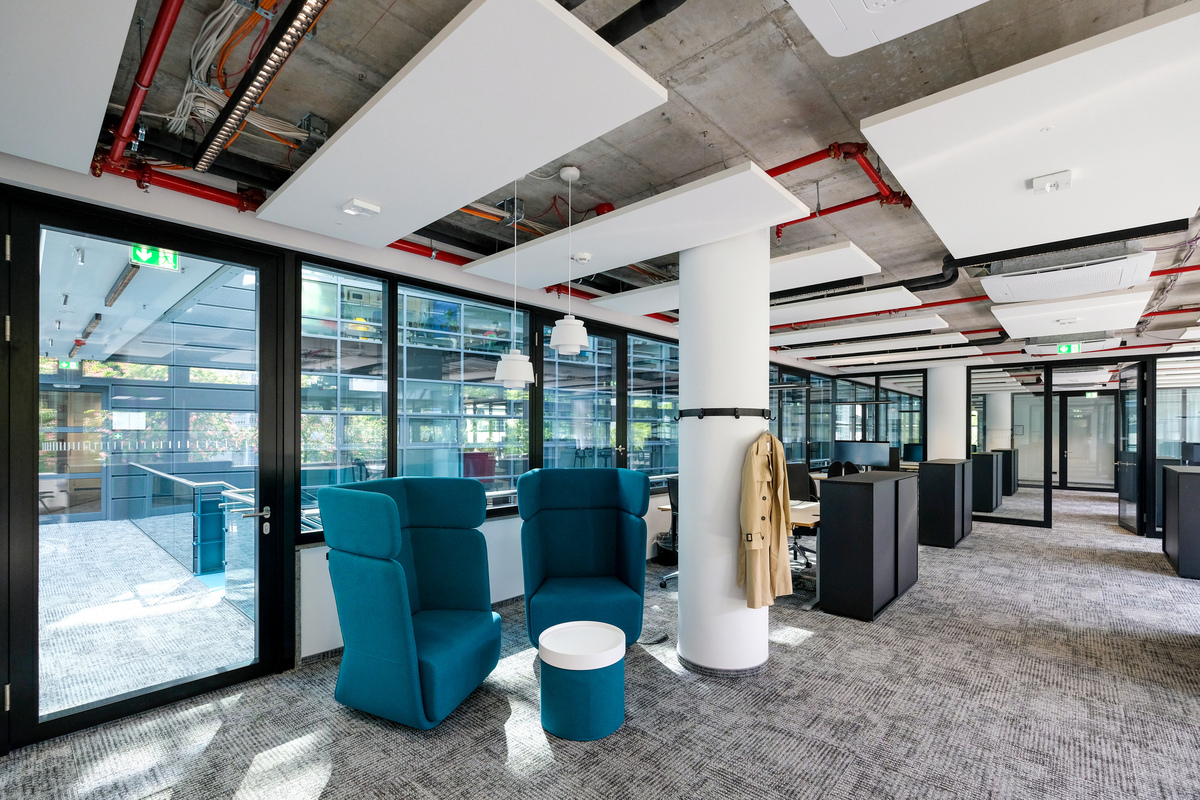 Modern open office with blue cushioned seating, a round white table, workstations, glass walls, and ceiling panels. A tan coat hangs on a column, and exposed pipes are visible on the ceiling. Natural light fills the space.