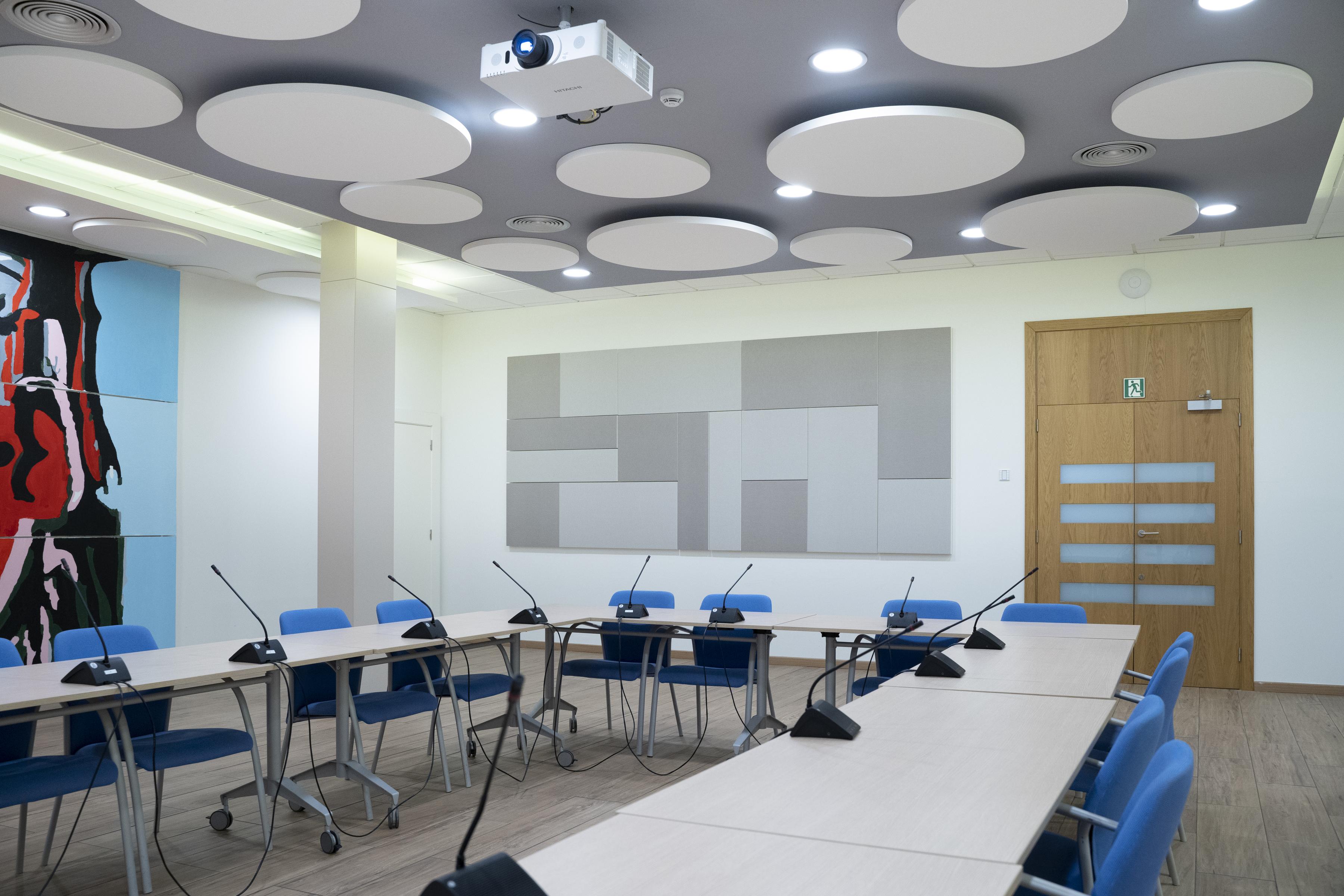 Large conference room with a U-shaped seating arrangement, grey acoustic wall panels in a pattern and white circle shaped free-hanging ceiling panels. 