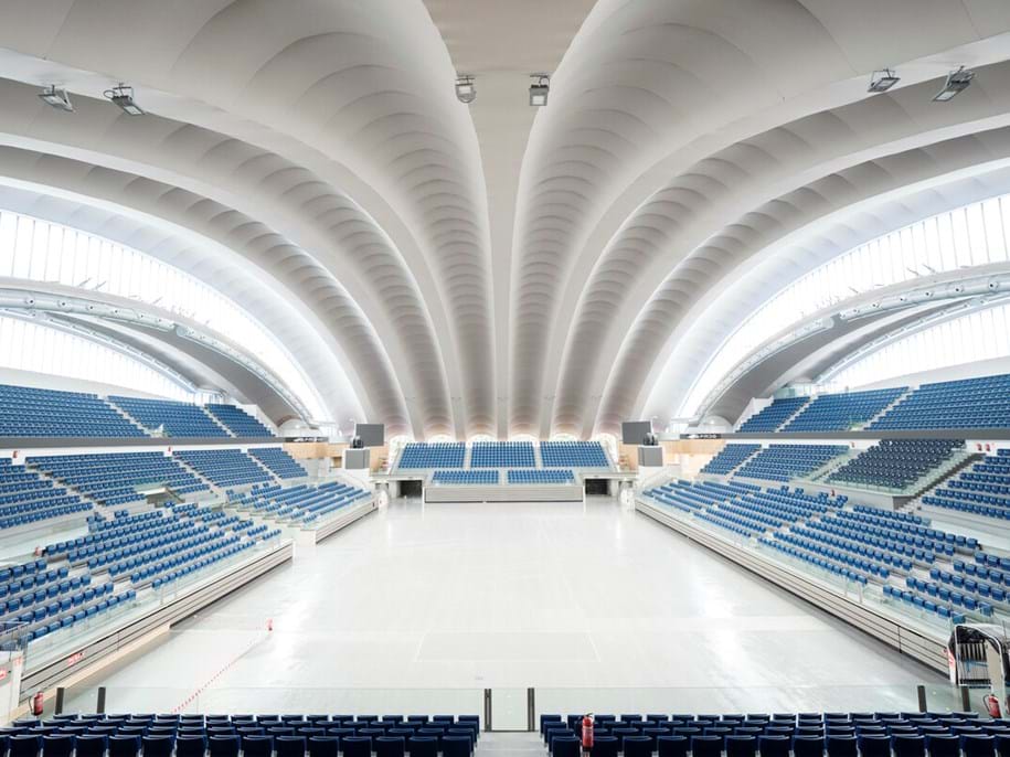 A spacious, modern indoor arena with blue seats, white arched ceiling panels, and abundant natural light from large windows curving along the roof. The arena floor is empty.