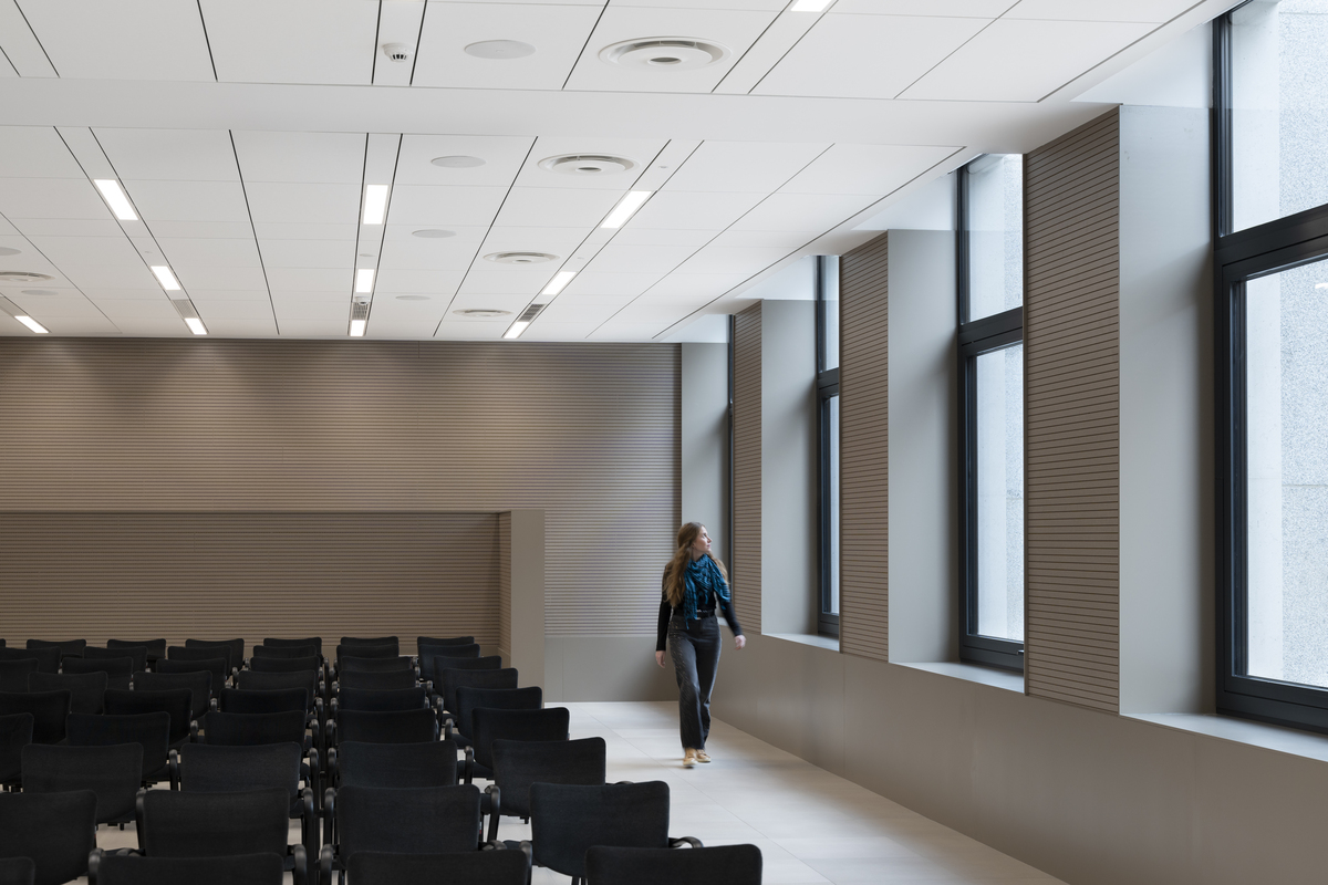 White, suspended acoustic ceiling in large conference room with beige floor and walls,  black chairs in multiple rows and a woman looking out the big windows