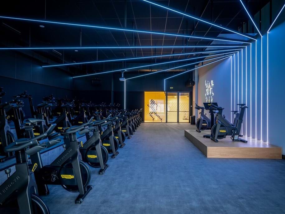 A modern indoor cycling studio with rows of stationary bikes, neon blue ceiling lights, a mirrored wall, and a small platform at the front. An illuminated doorway with a yellow glow is visible in the background.