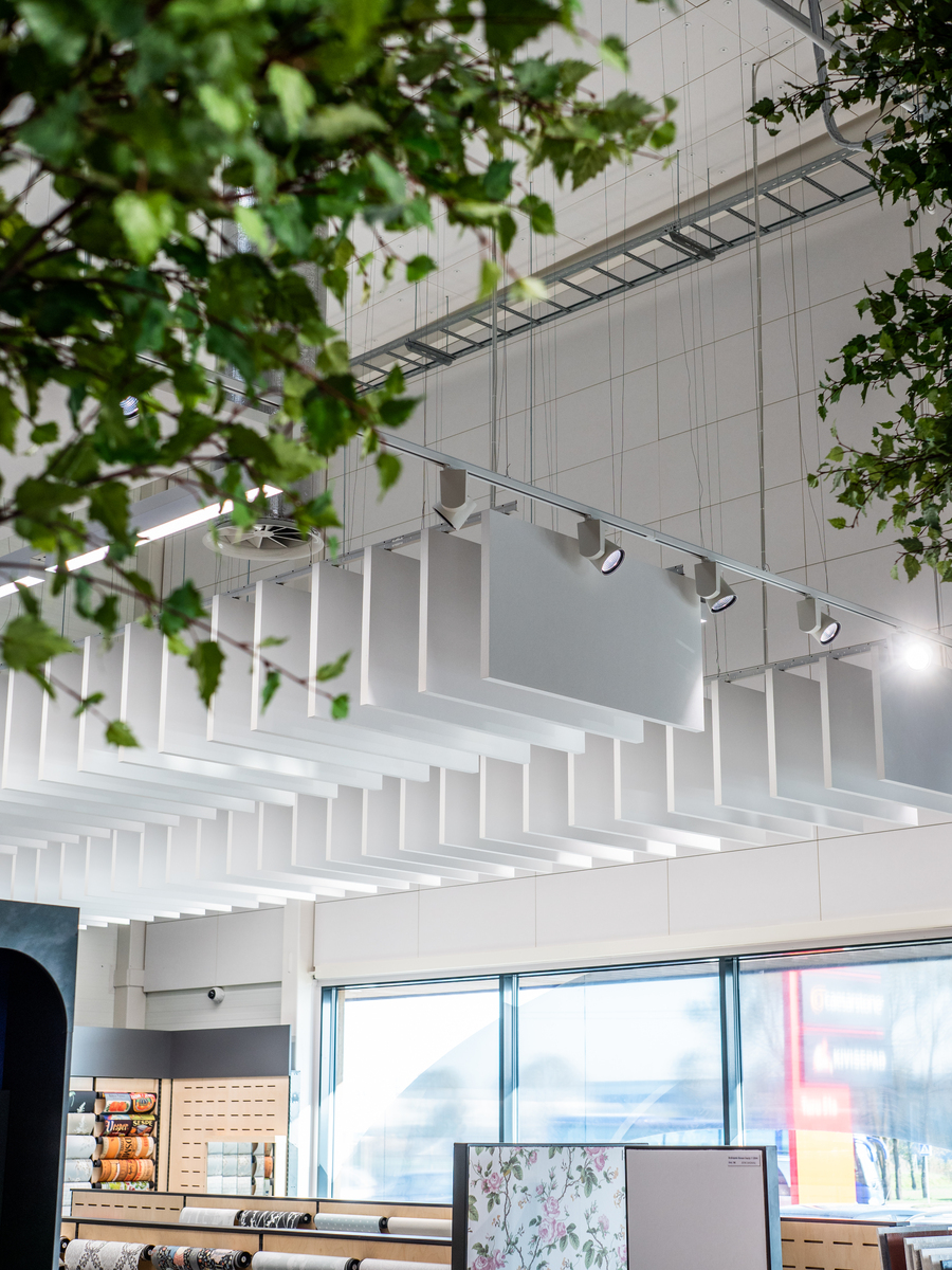 Modern interior of a shop with large windows, white ceiling panels, hanging lights, shelves displaying rolls of wallpaper, and leafy green branches partially visible in the foreground.
