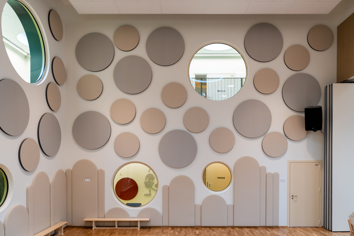 A modern room with a wooden floor, light-coloured walls decorated with large circular panels and round windows. Benches sit against the wall, and a small white door is on the right side. The decor is playful and geometric.