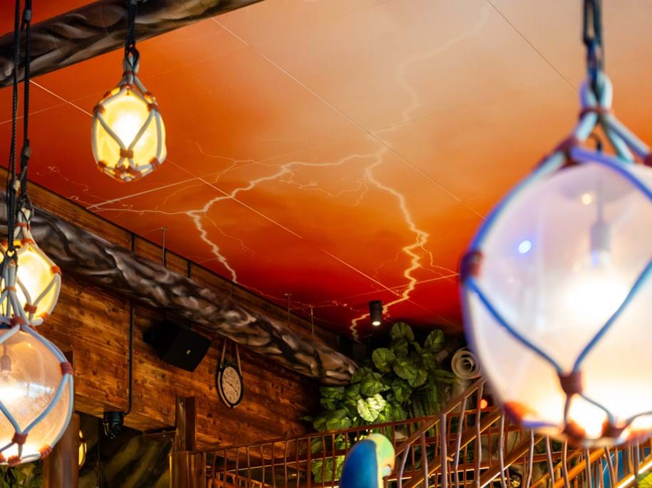 Hanging lanterns with glass globes and rope netting are suspended from a wooden ceiling with a painted lightning pattern, above a rustic interior with greenery and a clock on the wall.