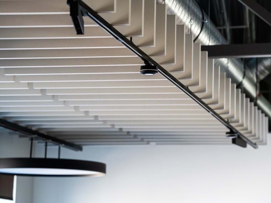 Angled view of a modern ceiling with evenly spaced, rectangular acoustic panels and exposed metal ductwork, featuring a circular light fitting hanging below.