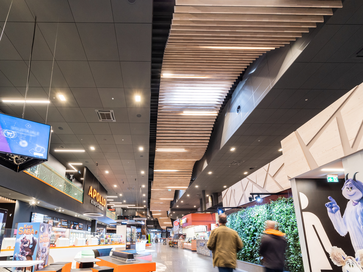 A modern cinema foyer with a striking wooden ceiling feature, bright lighting, ticket counters, film posters, a green plant wall, and two people walking through the spacious interior.
