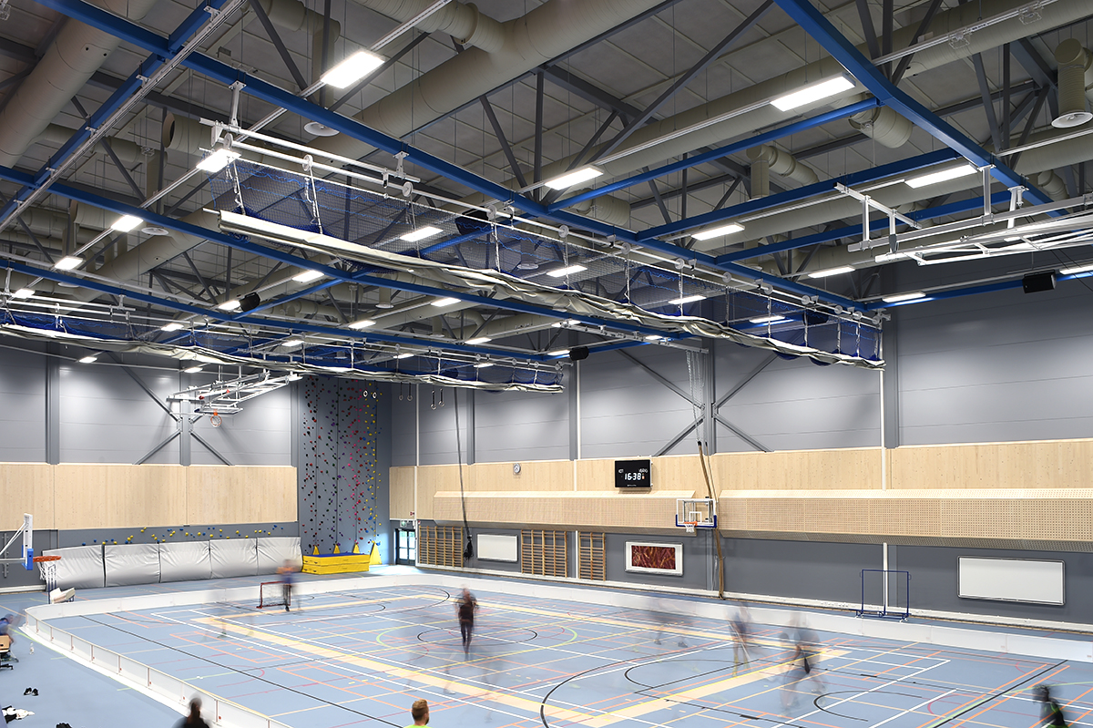 A spacious indoor sports hall with a high ceiling, a climbing wall, basketball hoops, and multiple courts. Several blurred figures are playing sports on the blue-marked floor. Bright lights illuminate the area.