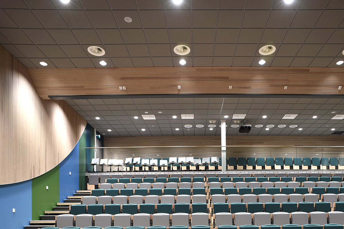 Modern lecture theatre with rows of empty grey and teal seats, tiered seating, wood-panelled walls, blue and green accent wall, and a ceiling with recessed lighting.