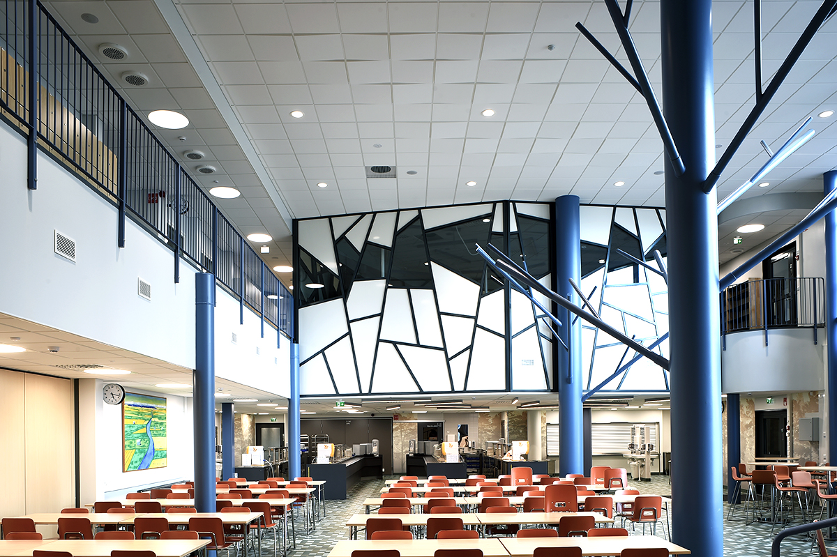 A modern canteen with orange chairs and white tables, high ceilings, blue columns, and a geometric black-and-white wall feature. The space is open, bright, and contemporary, with a first-floor railing.
