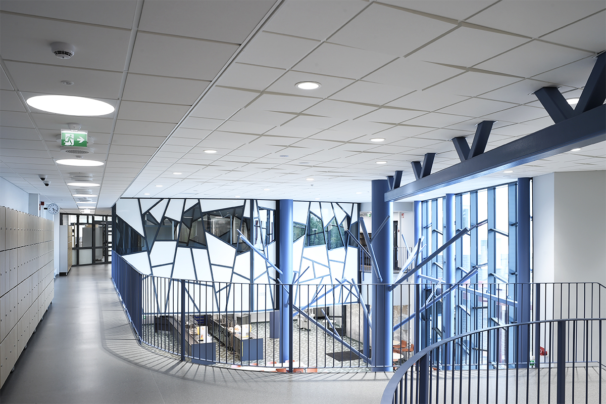 A modern building interior with geometric windows, blue columns, a metal railing, white lockers on the left, and an open, brightly lit corridor.