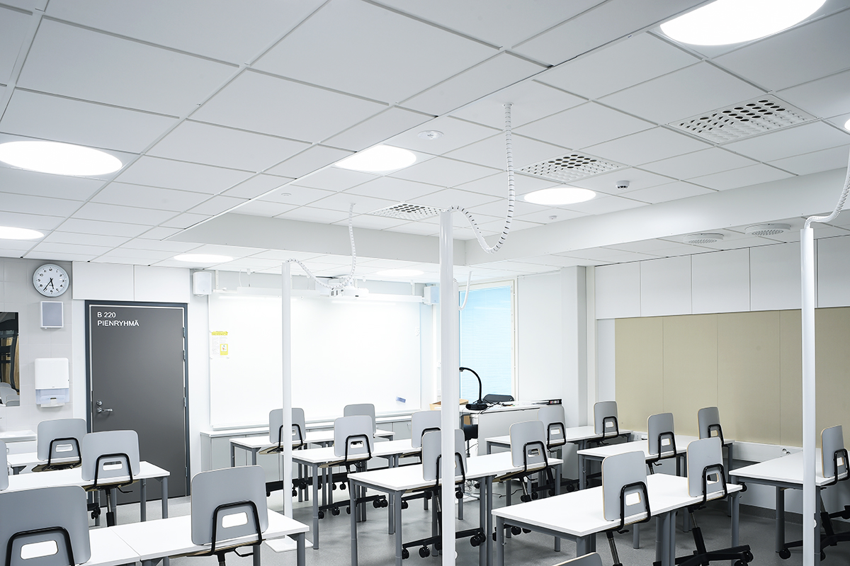 A modern, empty classroom with white walls, white desks and grey chairs, bright ceiling lights, a clock, and a whiteboard at the front. The room appears clean and organised, with large windows letting in light.