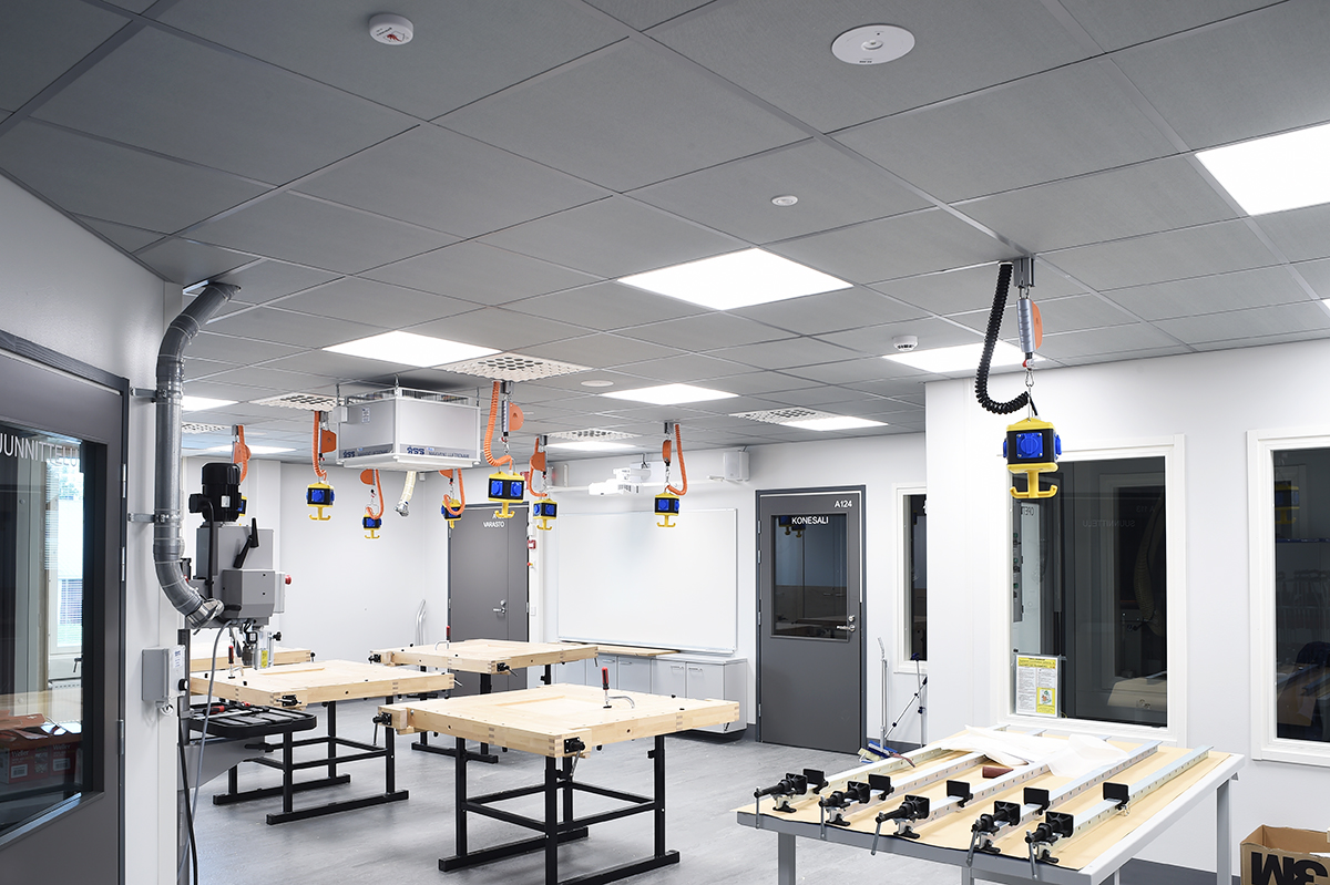 A modern workshop with multiple workbenches, ceiling-mounted equipment, and various tools. The room is clean, brightly lit, and equipped with advanced ventilation and technology. No people are present.