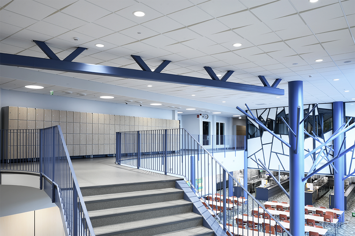 A modern, brightly lit interior with blue beams, a staircase, metal railings, lockers along the wall, and tables with chairs in an open area below. Large windows let in natural light.