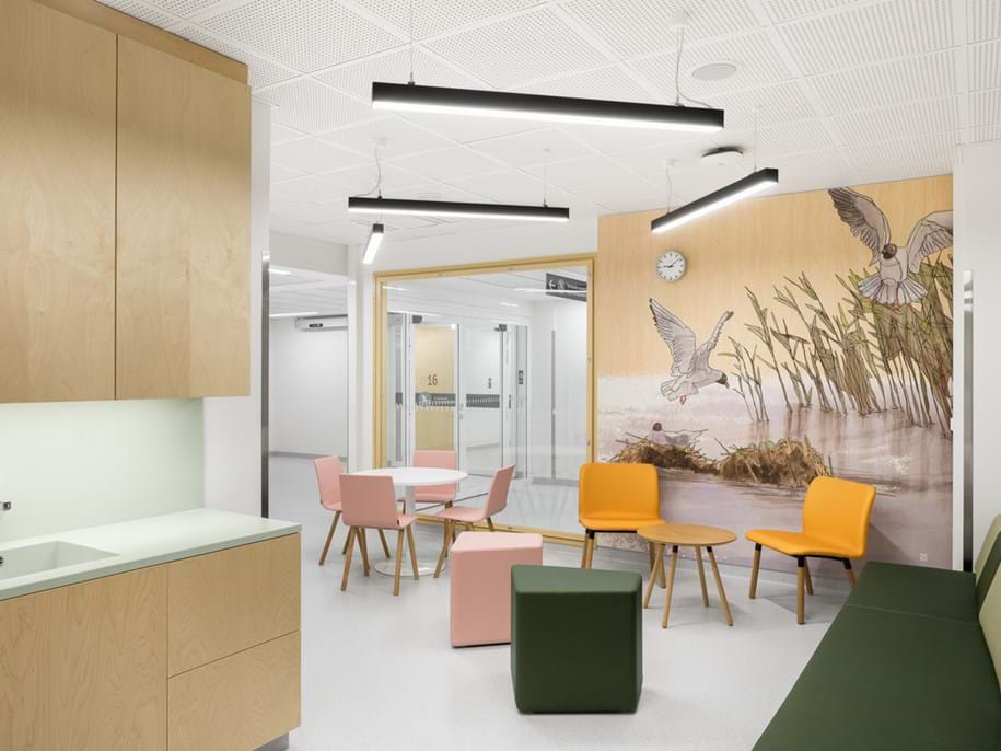 A bright, modern waiting area with pastel chairs, small tables, and green benches. A wall mural shows birds by water and reeds. Light wood cupboards and white walls create a clean, calming atmosphere.