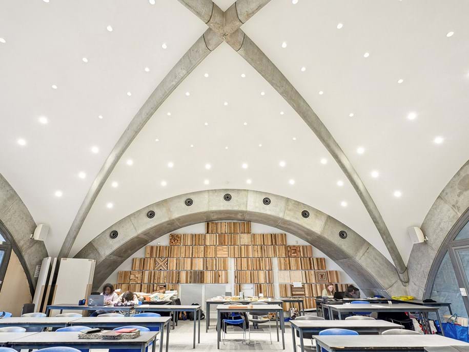 A modern classroom with arched concrete ceilings, bright overhead lights, shelves filled with wooden samples, and students working at desks. The space appears spacious and contemporary.