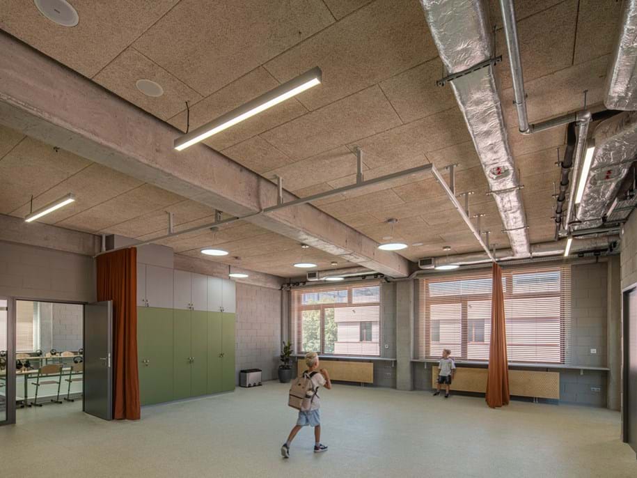 A spacious, modern classroom with exposed ceilings, large windows, green cupboards, and two children wearing rucksacks walking across the room. The space has natural light and visible ductwork.