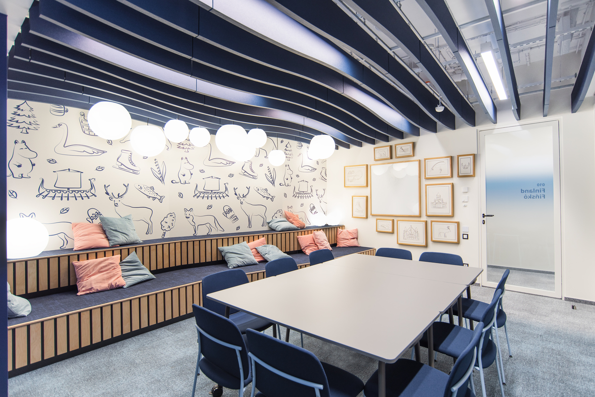A modern meeting room with a large table surrounded by blue chairs. The room has a wall with playful animal and nature drawings, framed sketches, and tiered bench seating with colourful pillows. Round ceiling lights add to the ambiance.