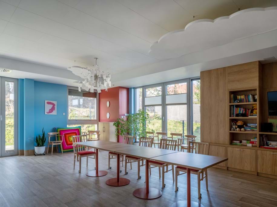 A bright, modern room with large windows, wooden tables and chairs, colourful walls, bookshelves, plants, playful decor, and cloud-shaped ceiling panels, filled with natural light.