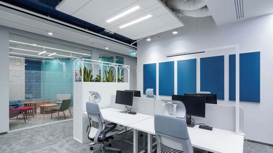 Ein moderner Büroraum mit weißen Schreibtischen und hellblauen Bürostühlen, die jeweils mit Computern und Monitoren ausgestattet sind. Blaue Akustikpaneele zieren die Wand. Im Hintergrund befindet sich ein gläserner Besprechungsraum mit bunten Stühlen und einem gemusterten Teppich.