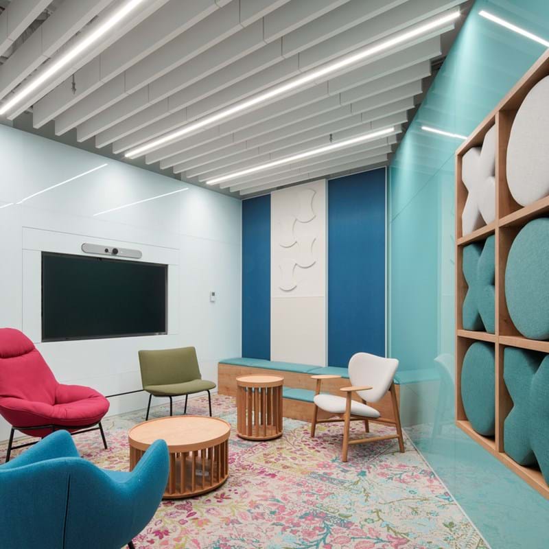 A modern office lounge with colourful, eclectic furniture. There are red, blue and white chairs, wooden tables, a patterned rug, and a wall with soundproof panels. The ceiling has white acoustic beams and linear lighting.